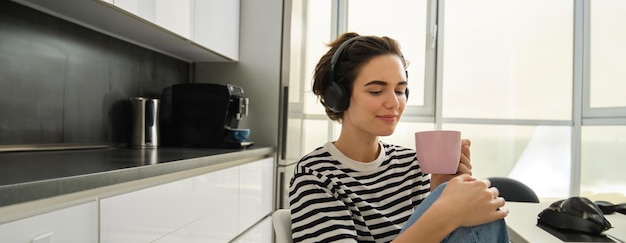Photo gratuite portrait d'une femme souriante et détendue buvant du thé chaud et écoutant de la musique ou un livre électronique dans la cuisine