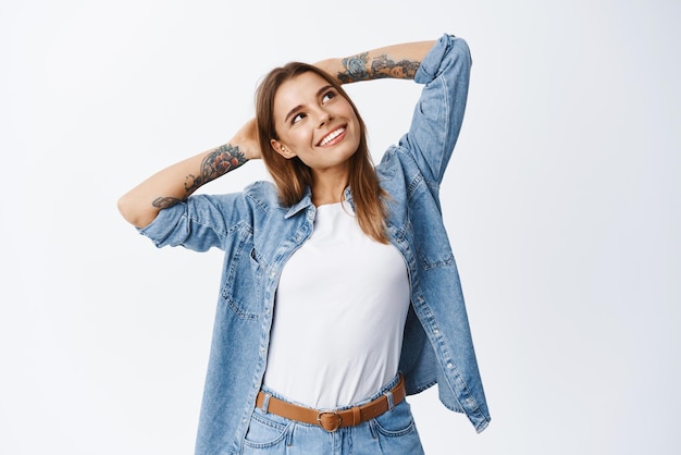 Portrait d'une femme souriante et détendue, tenant les mains derrière la tête, regardant le coin supérieur droit, rêveuse, se reposer, avoir du temps libre, debout sur fond blanc