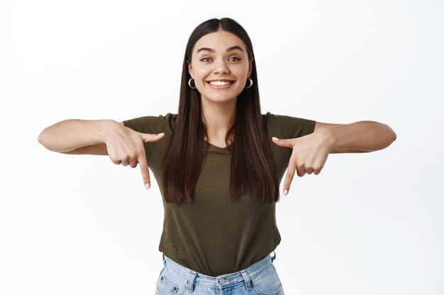 Portrait d'une femme souriante enthousiaste montrant une publicité en bas, pointant les doigts vers le bas et l'air heureux à l'avant, debout sur un mur blanc