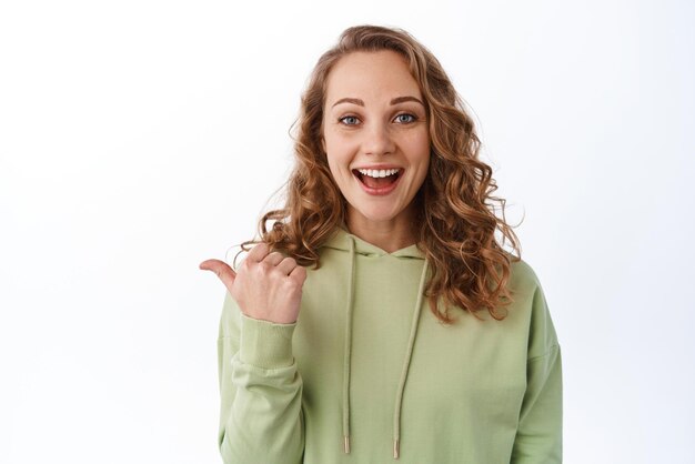 Portrait d'une femme souriante joyeuse avec des cheveux blonds bouclés pointant le pouce vers la gauche montrant l'espace de copie de côté regarde étonné démontrer un lien ou un logo sur fond blanc