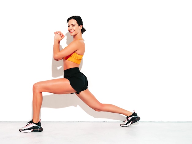 Portrait de femme souriante de remise en forme en vêtements de sport noirs Sexy jeune beau modèle avec un corps parfait Femme isolée sur un mur blanc en studio S'étirer avant l'entraînement Faire des fentes