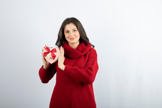 Portrait d'une femme souriante tenant dans les mains une boîte cadeau en forme de coeur sur un mur blanc.