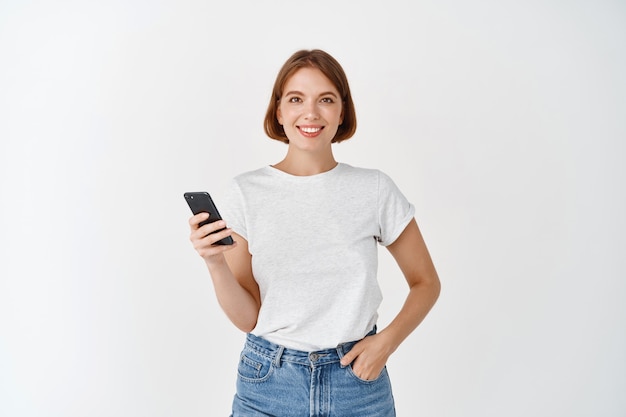 Portrait d'une femme souriante utilisant un smartphone, discutant sur les réseaux sociaux, debout avec un téléphone portable contre un mur blanc