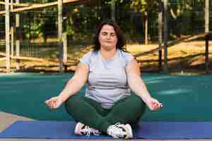 Photo gratuite portrait d'une femme en surpoids en combinaison de sport méditant sur un tapis de yoga sur un terrain de sport