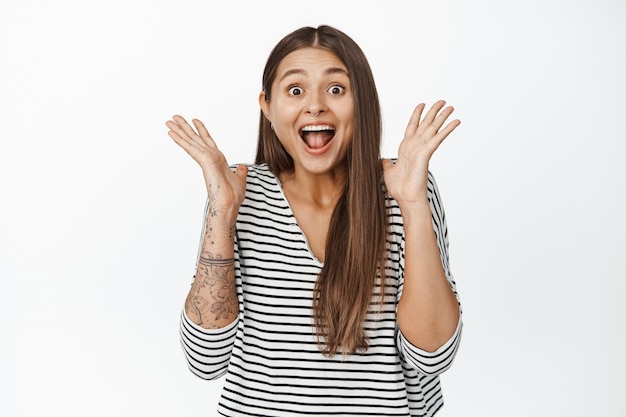 Portrait d'une femme surprise et heureuse réagit aux grandes nouvelles, serrant la main près du visage et criant de joie sur blanc