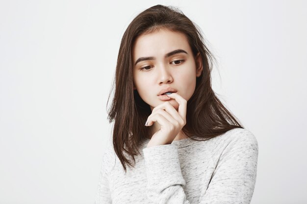 Portrait d'une femme tendre rêveuse avec une expression réfléchie, tenant la main près de la bouche tout en mordant le doigt et regardant vers le bas