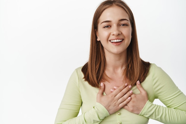 Portrait d'une femme touchée dire merci, tenir la main sur le cœur et sourire heureux, recevoir des éloges, un cadeau ou un compliment, debout contre un mur blanc