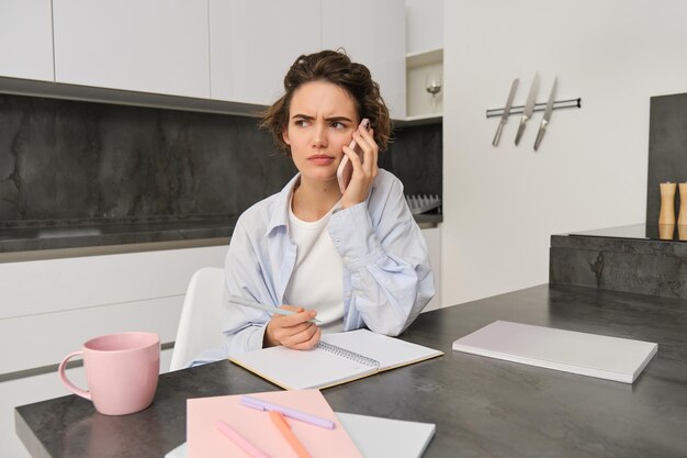 Portrait d'une femme travaillant à domicile appelant quelqu'un sur un téléphone portable et ayant l'air confuse tout en la tenant