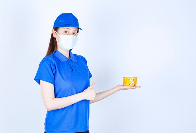 Portrait de femme en uniforme et masque médical avec gobelet en plastique montrant le pouce vers le haut