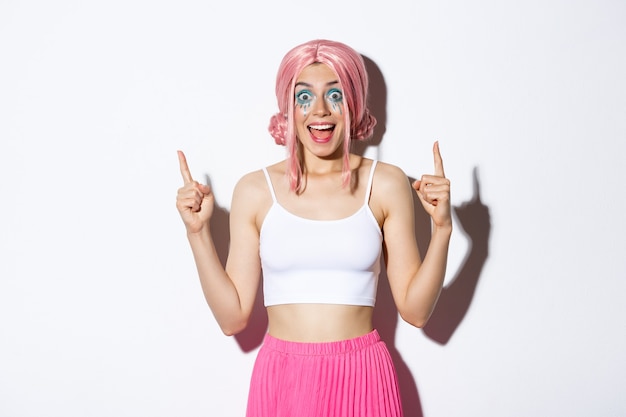 Portrait d'une fêtarde excitée avec une perruque rose et un maquillage d'halloween, l'air étonné tout en pointant les doigts vers le haut, debout sur fond blanc