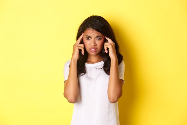 Portrait d'une fille afro-américaine frustrée, fronçant les sourcils et touchant la tête, l'air affligé devant la caméra, debout sur fond jaune.