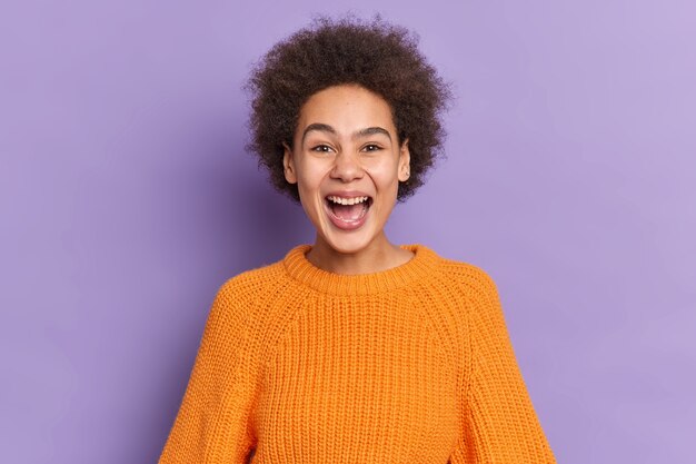 Portrait de fille afro-américaine positive rit joyeusement garde la bouche ouverte a des dents blanches entend des nouvelles drôles habillé en pull tricoté orange.