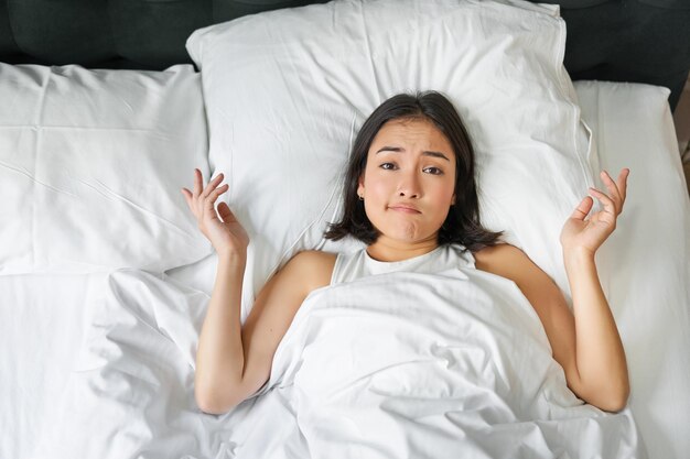 Portrait d'une fille asiatique allongée dans son lit et haussant les épaules semble perplexe avant de s'endormir