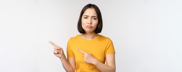 Portrait d'une fille asiatique brune déçue pointant les doigts à gauche en grimaçant contrarié montrant qch désagréable debout sur fond blanc