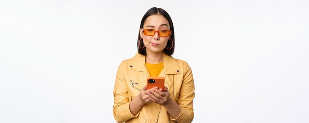 Portrait d'une fille asiatique confuse dans des lunettes de soleil tenant un smartphone à l'aide d'un téléphone portable et regardant puzz