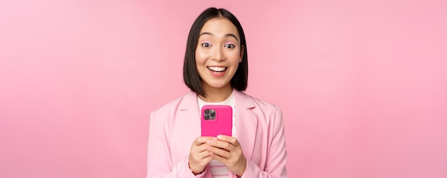 Portrait d'une fille asiatique en costume avec un smartphone souriant et heureux debout sur fond de studio rose