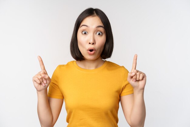 Portrait d'une fille asiatique impressionnée pointant les doigts vers le haut dire wow à la recherche de l'appareil photo étonné debout sur fond blanc