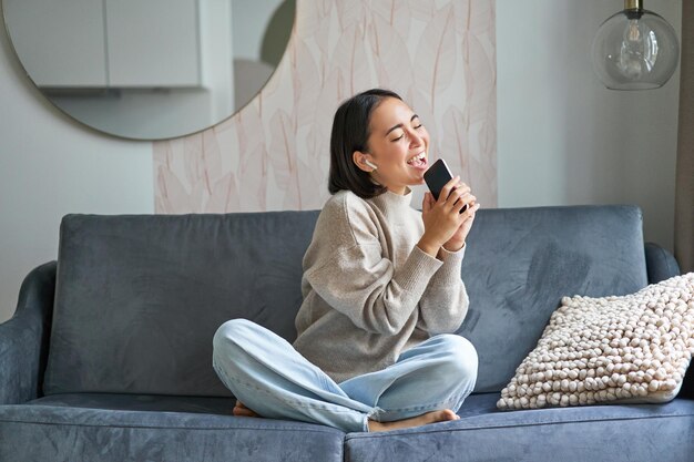 Portrait d'une fille asiatique insouciante assise à la maison écoute de la musique dans des écouteurs et chante sur un smartphone