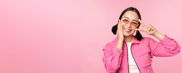 Portrait d'une fille asiatique kawaii à lunettes de soleil montrant la paix vsign près des yeux et sourit mignon à la caméra posant dans des vêtements à la mode sur fond rose