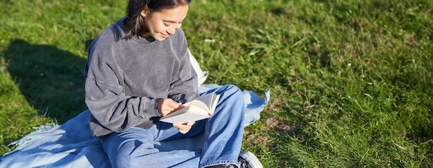 Photo gratuite portrait d'une fille asiatique lisant un livre assise sur sa couverture dans un parc avec de l'herbe verte souriant heureusement