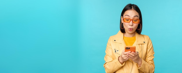 Portrait d'une fille asiatique à lunettes de soleil regardant l'écran du smartphone avec une expression de visage étonné impressionné wow face debout sur fond bleu