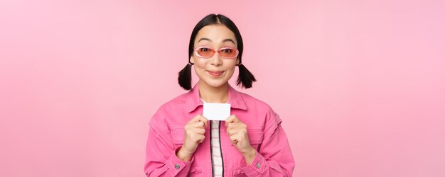 Portrait d'une fille asiatique moderne et élégante montre une carte de crédit à prix réduit et a l'air heureux de payer le concept sans contact de shopping debout sur l'espace de copie de fond rose