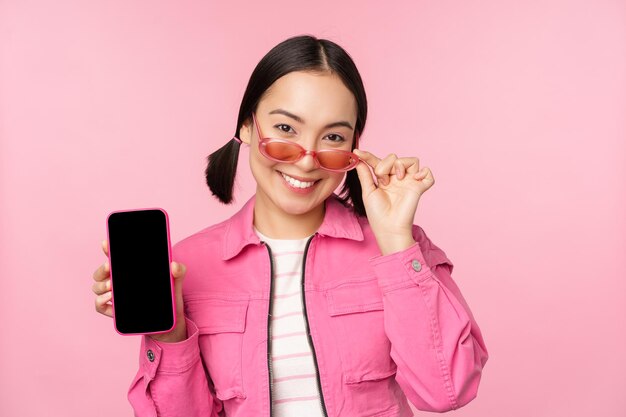 Portrait d'une fille asiatique moderne et élégante montre une interface d'application pour smartphone à écran de téléphone portable debout dans des lunettes de soleil sur fond rose