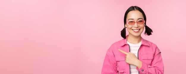 Portrait d'une fille asiatique souriante dans des lunettes de soleil tenues élégantes pointant le doigt vers la gauche montrant une bannière publicitaire debout sur fond rose Copier l'espace