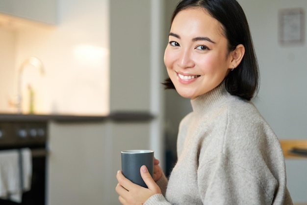 Portrait d'une fille asiatique souriante avec une tasse de café, boire du thé chaud et se réchauffer à la maison