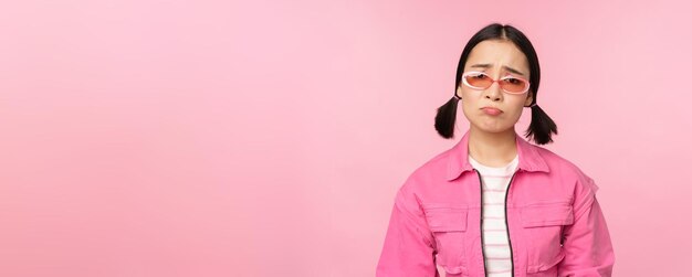 Portrait d'une fille asiatique triste et sombre boudant de déception debout bouleversée sur fond de studio rose