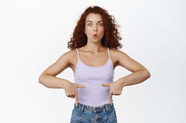 Portrait d'une fille au gingembre aux cheveux bouclés surprise, une femme haletant et pointant vers le bas avec une expression de visage étonnée et excitée, montrant une publicité, fond blanc