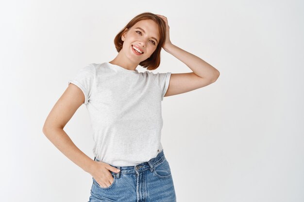 Portrait d'une fille de beauté naturelle avec un maquillage léger, touchant les cheveux courts et souriante heureuse, debout en t-shirt décontracté avec un jean contre un mur blanc