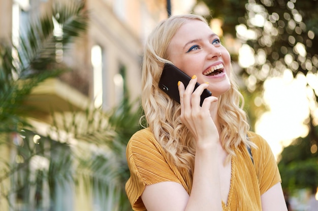 Portrait d'une fille blonde assez joyeuse regardant joyeusement au loin tout en parlant sur un téléphone portable dans la rue de la ville