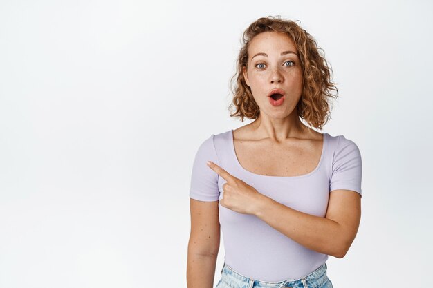 Portrait d'une fille blonde bouclée à la surprise, pointant le doigt vers la gauche avec une expression de visage intriguée sur blanc.