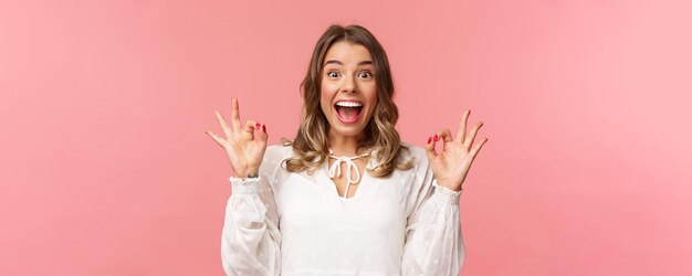 Portrait d'une fille blonde gaie excitée et optimiste en robe blanche dansant et montrant des signes corrects avec un sourire rayonnant heureux excité par le taux du festival de musique film génial recommander produit