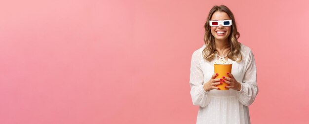 Portrait d'une fille blonde heureuse et insouciante en robe blanche appréciant de regarder un film en 3d portant des lunettes mangeant du pop-corn et souriant assister au cinéma sur fond rose