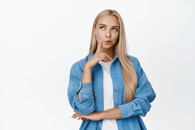 Portrait d'une fille blonde idiote pensant, regardant de côté pensif, debout sur du blanc, une femme réfléchit à quelque chose