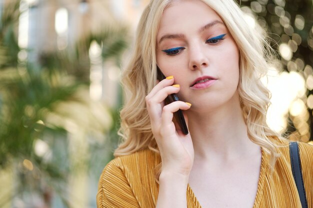 Portrait d'une fille blonde sérieuse avec du maquillage parlant pensivement sur un téléphone portable en plein air seul