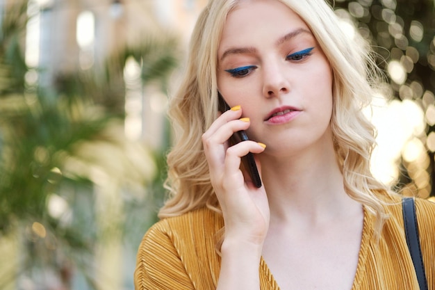 Portrait d'une fille blonde sérieuse avec du maquillage parlant pensivement sur un téléphone portable en plein air seul