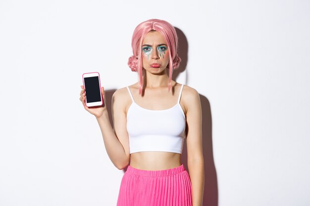 Portrait de fille bouder se plaindre et montrant quelque chose de décevant sur l'écran du téléphone mobile, debout dans les cheveux roses et tenue de fête.