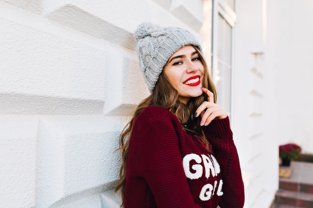 Portrait fille brune aux cheveux longs en pull d'hiver sur un mur gris dans la rue. Elle porte un bonnet tricoté, touchant le visage, souriant.