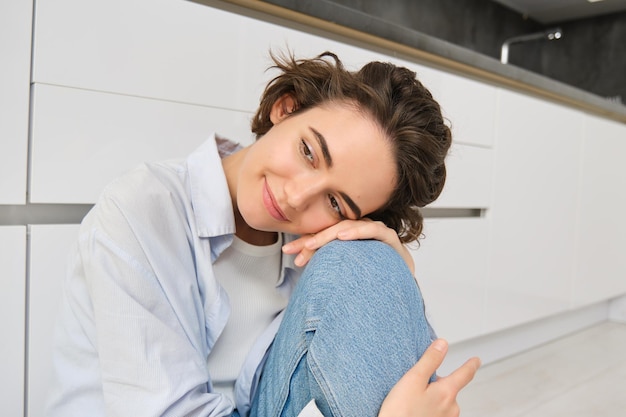 Portrait d'une fille brune rêvant éveillée assise sur le sol de la cuisine à la maison et souriante regardant de côté l'esprit