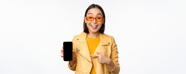 Portrait d'une fille coréenne élégante dans des lunettes de soleil souriant pointant du doigt l'écran du smartphone montrant m