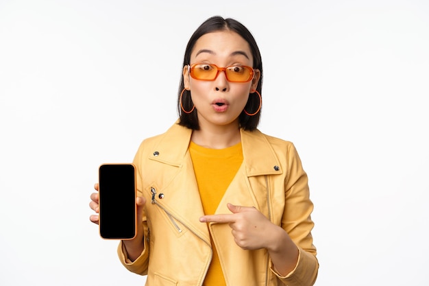 Portrait d'une fille coréenne élégante à lunettes de soleil souriant pointant du doigt l'écran du smartphone montrant l'application de téléphonie mobile debout sur fond blanc