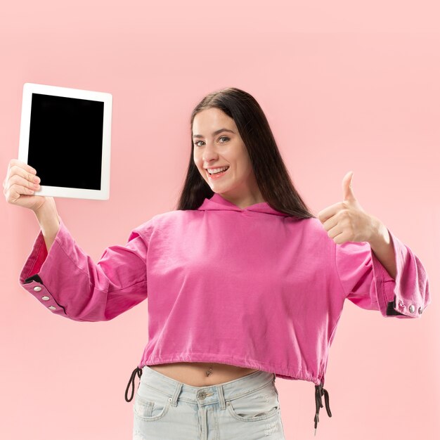 Portrait d'une fille décontractée confiante montrant un écran vide d'une tablette isolée sur fond rose.