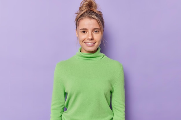 Portrait d'une fille du millénaire satisfaite qui sourit doucement a une peau saine pas de maquillage beauté naturelle regarde directement la caméra avec une expression satisfaite porte un poloneck vert isolé sur fond violet