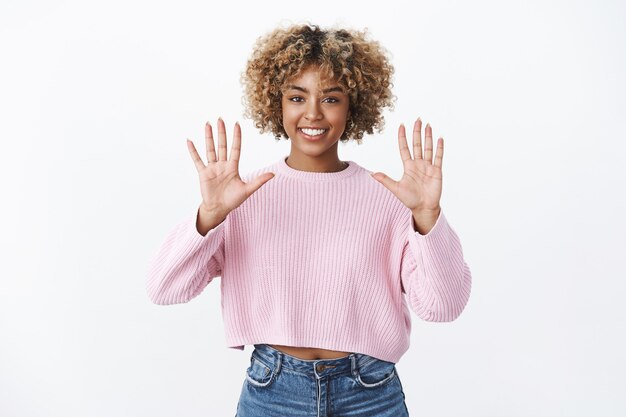 Portrait d'une fille élégante afro-américaine optimiste et joyeuse avec une coupe de cheveux afro blonde souriante ravie et confiante en levant les mains et en montrant le numéro dix, posant contre un mur blanc