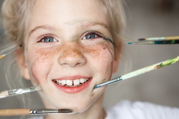 Portrait de fille européenne blonde ludique avec des taches de rousseur et de la peinture sur son visage, jouant avec des pinceaux. Fille blonde assez joyeuse, souriant avec des dents.