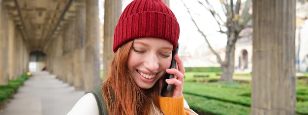 Portrait d'une fille européenne rousse au chapeau rouge passe un appel téléphonique, se promène en ville et parle à un ami