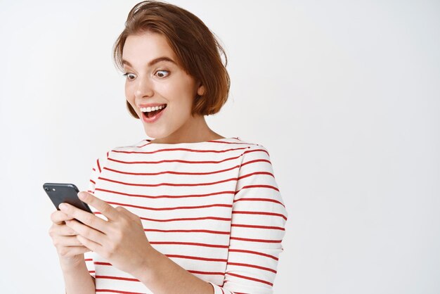 Portrait d'une fille excitée à la surprise de l'écran du smartphone lisant de grandes nouvelles debout heureux sur fond blanc